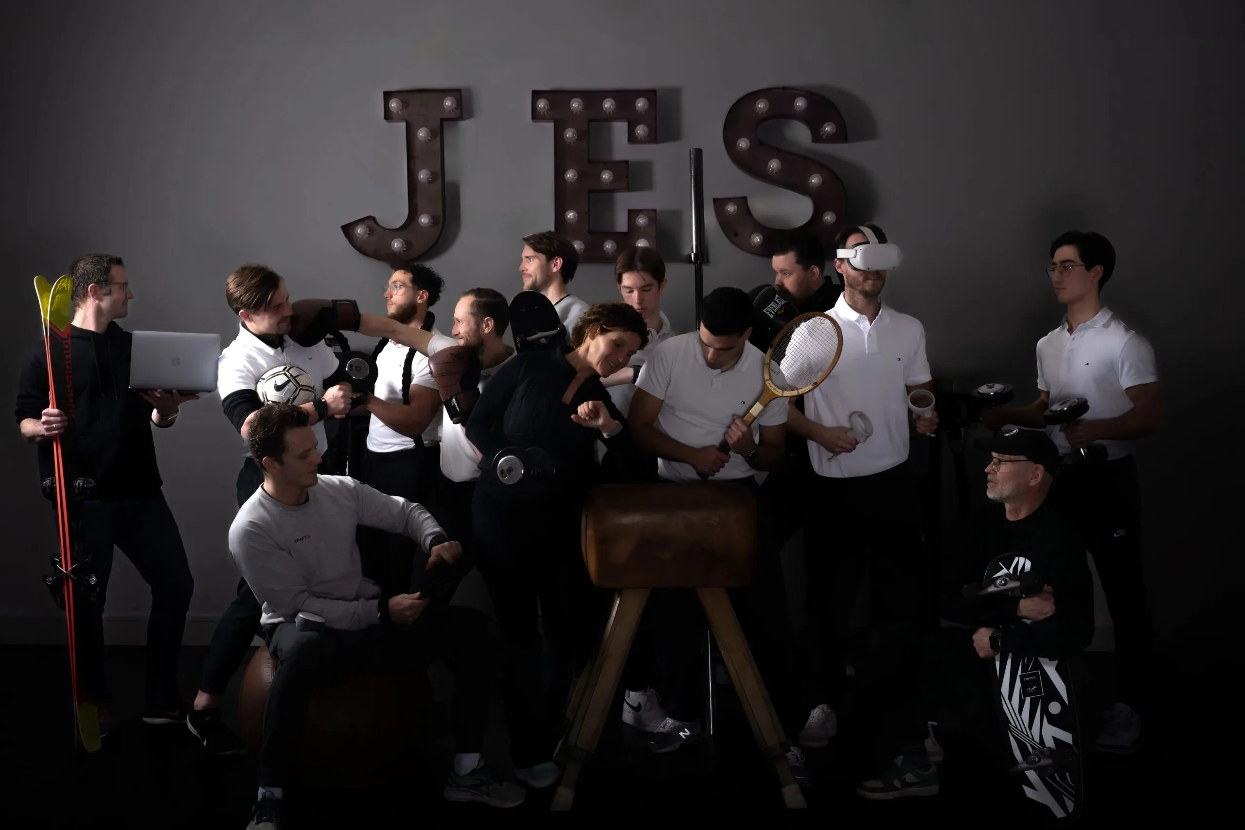 Het hele team van JES poseert met ski’s, een bokshandschoen, een tennisracket en een bok onder de JES-lichtletters